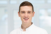 Portraitbild von Caelán Haney-Aubert: Mann mit brauen Haaren im weißen Arzt-Kittel