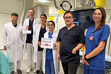 Team der Kardiologie der UMM in einem Herzkatheterlabor. Eine Mitarbeiterin hält eine Übersicht zur Studie, ein Kollege ein Modell mit der Aufschrift „Frauenherzen“.